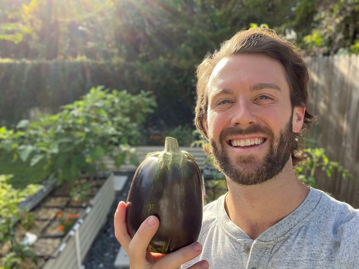 Man holding a large eggplant in front of a 17" Tall, 10-In-1 Metal Raised Garden Bed