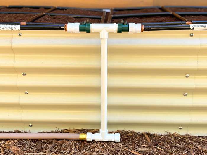 Double Style Garden Grid Manifold standing infront of an ivory metal raised garden bed with two Garden Grid watering systems connected to the top of the manifold.